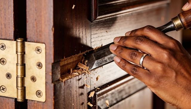 Close-up hand chiseling door edge with bench chisel, wood shavings and brass hinge