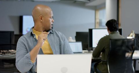 Male coworkers collaborating in open office, one turning and holding glasses by laptop