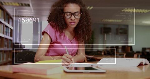 Focused student writing notes at library desk with tablet and futuristic data hud overlay