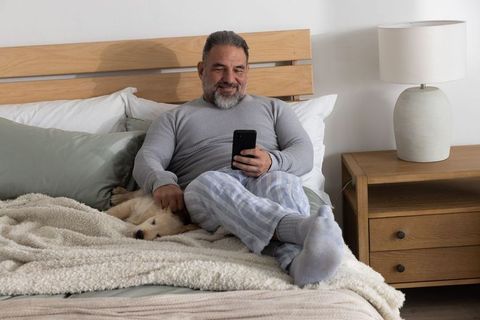 Mature man relaxing in bed using smartphone and petting golden retriever in striped pajamas
