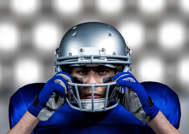 Depicts an American football player wearing blue jersey and protective gear, adjusting his helmet with a serious expression. Ideal for use in sports-related content, motivational materials highlighting determination and focus, promotional materials for sporting events, or articles about team sports and athleticism.