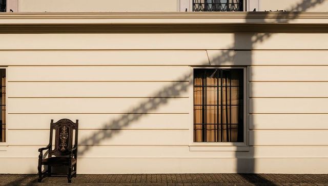 Ornate wooden armchair casting long shadow on cream heritage facade with barred window