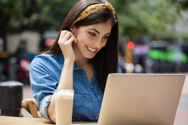 Young woman sitting at a sidewalk cafe in an urban setting, smiling while using a laptop. She is wearing a casual denim shirt and a headband, with a glass of iced coffee on the table. Ideal for concepts related to remote work, freelancing, modern lifestyle, technology, and urban living.