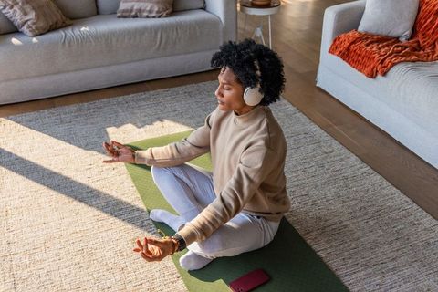 African american woman meditating with headphones on green yoga mat in sunlit living room