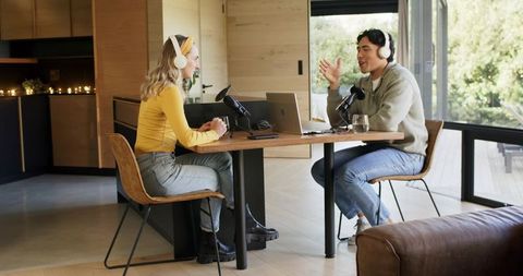 Diverse podcasters recording at home on wooden table with laptop, microphones, headphones