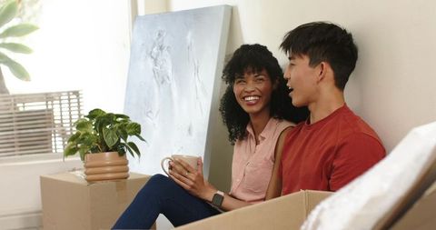 Diverse couple unpacking in sunlit new home smiling while holding mug and smartwatch