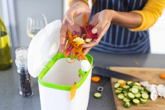 Woman disposing vegetable peels into a kitchen compost bin while preparing food. Ideal for illustrating sustainable living, eco-friendly practices, and home cooking. Useful for articles on waste management, green living, and kitchen organization.