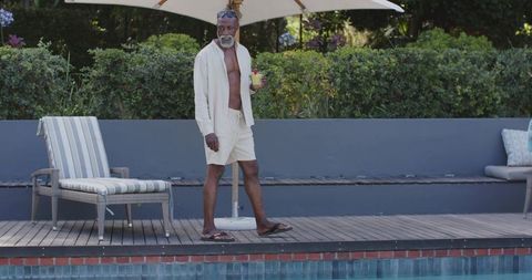 Mature african man strolling poolside holding cocktail in light resort shirt and shorts