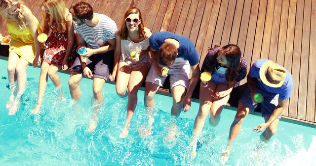 Group of friends sitting on poolside deck with feet in water, laughing and enjoying each other's company. They are holding colorful drinks, having a great time in outdoor summer setting. Use for summer vacation, friendship, leisure time, and outdoor lifestyle concepts.