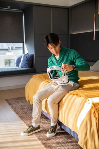 Asian man inspecting white vr helmet while sitting on bed in sunlit modern bedroom