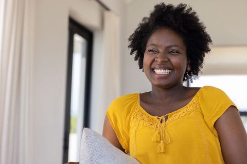 African american woman smiling and holding cushion on sofa in mustard top, sunlit interior