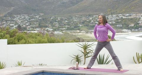 Middle-aged woman practicing wide-legged yoga pose on coastal terrace beside pool