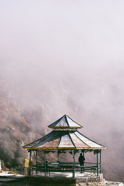 Image shows a person standing in a rustic wooden gazebo, looking out at a misty, mountainous landscape. Ideal for use in travel websites, nature blogs, or inspirational posters conveying solitude and tranquility. Perfect for illustrating peaceful outdoor settings or adding a serene ambiance to nature-related content.