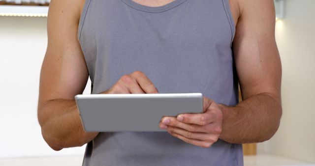 Man in a grey tank top using a tablet indoors. Ideal for depicting everyday technology use, home leisure, remote work, online communication, or browsing the internet activities.