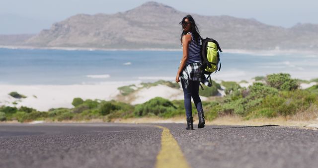 African american woman with backpack trying to hitch a lift while walking on road. road trip travel and adventure concept