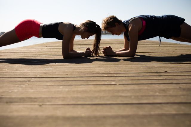 Side view of two Caucasian female rowers wearing sportswear and training on a jetty by the river, exercising doing planks with river and blue sky in the background.