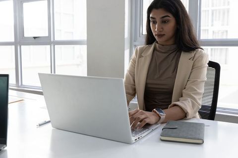 Indian businesswoman working on laptop in modern minimalist sunlit office with notebook