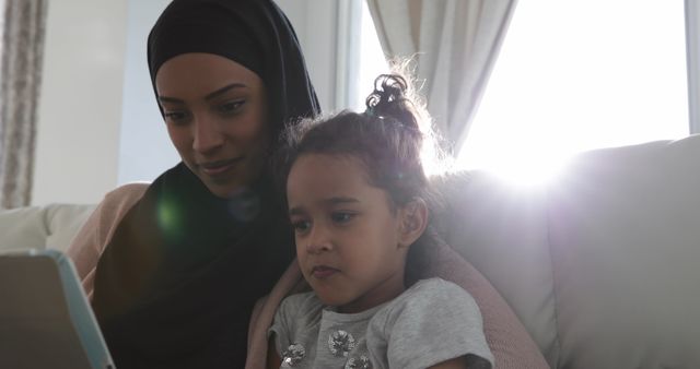 The image shows a mother and daughter sitting on a couch using a tablet together in a bright, cozy living room. The mother is wearing a hijab, indicating an Islamic cultural context. This can be used to represent family bonding, digital learning, parental involvement in education, or multicultural family life. Ideal for use in advertisements, educational materials, or family-oriented publications.