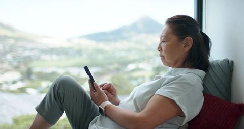 Senior asian woman relaxing on window seat using smartphone and smartwatch with valley view