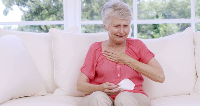 Elderly woman sitting on couch holding her chest and tissue, appearing in pain. Useful in articles about senior health, chest pain, cardiac issues, and emergency medical situations. Ideal for illustrating symptoms of health issues in the elderly and promoting awareness of senior health care.