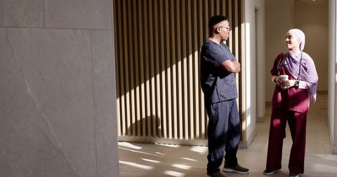 African american nurse and hijab-wearing clinician conversing in sunlit hospital corridor