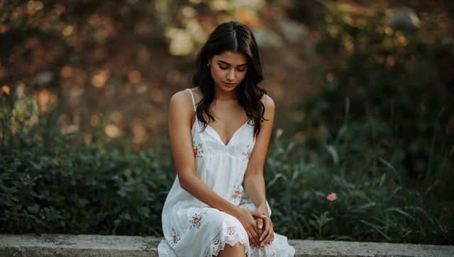 Young woman in white floral sundress sitting on garden ledge looking down