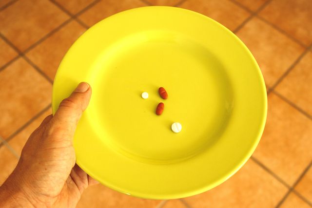 Shows a hand holding a yellow plate with a few pills and tablets on it, over a tiled floor. Useful for illustrating concepts related to healthcare, medication, daily vitamins and minimalism. Could be used in articles talking about medication schedules, health routines, and wellness topics.