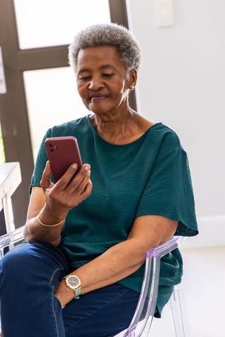 Senior african woman sitting on acrylic chair using smartphone at home, browsing and smiling