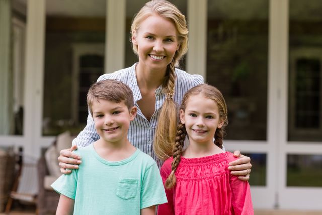 Mother standing with her two children outside their home, all smiling at the camera. Perfect for use in family-oriented advertisements, parenting blogs, and lifestyle magazines. Highlights themes of family bonding, happiness, and outdoor activities.