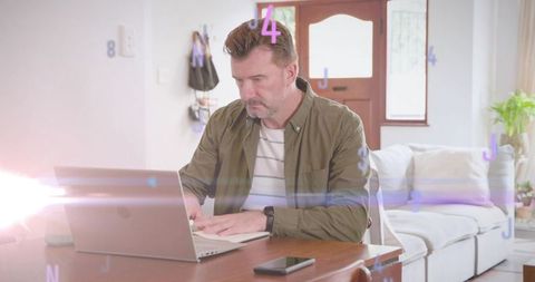 Mature man working remotely on laptop at home with smartphone, sunlit lens flare interior