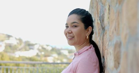 Smiling mid-adult woman leaning on stone wall on sunny terrace wearing pink polo and gold hoop earri