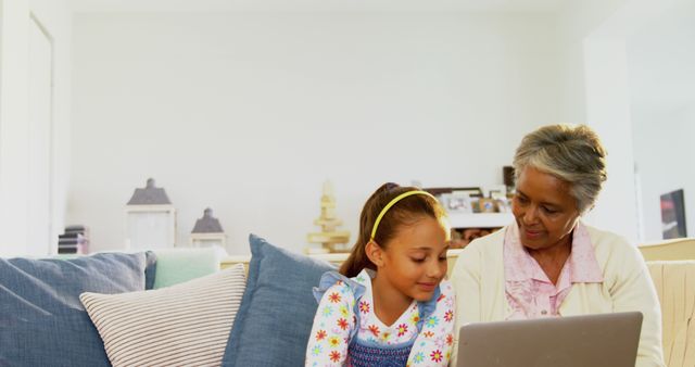 Grandmother and granddaughter are sitting on a comfortable sofa in a living room, using a laptop together. This image can be used to depict themes of family bonding, technology education, multi-generational relationships, and shared learning experiences at home. It is ideal for for home environment promotions, articles on family relationships, and educational content aimed at multiple age groups.