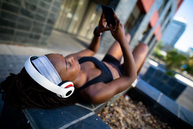 Fit african american woman wearing headphones, resting, using smartphone in street. healthy active lifestyle and outdoor fitness.