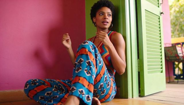 Sitting woman leaning on green louvered door barefoot on tiled floor in colorful patterned outfit