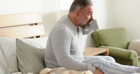 Mature man relaxing in morning drinking coffee on bed wearing striped pajamas with dog