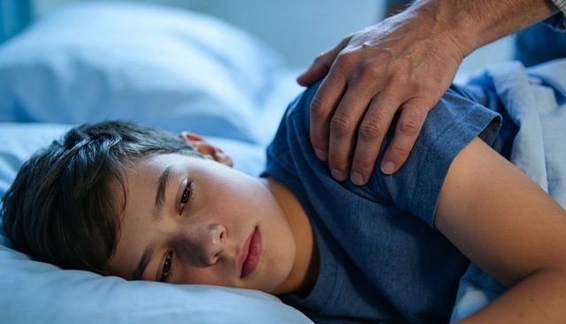 Preteen boy lying in bed receiving comforting hand on shoulder, feeling sad and soothed