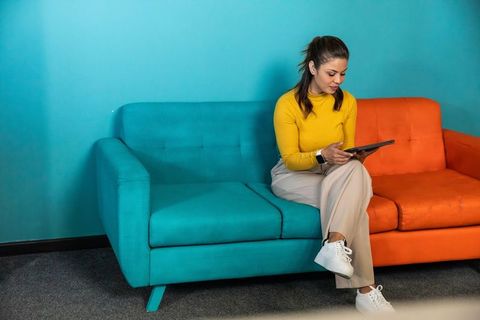Professional woman sitting between teal and orange sofas using tablet in modern lounge
