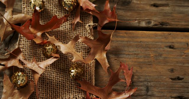 Rustic fall-themed decorations featuring dry leaves spread on burlap with golden accents on a wooden table. Ideal for seasonal marketing materials, crafting project inspiration, social media posts, and festive backdrop designs during autumn.