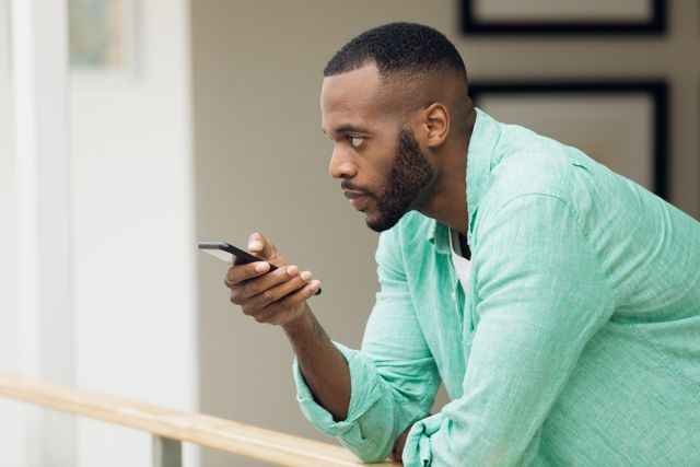 African-American man standing indoors, using a smartphone with a focused expression. He is leaning against a wooden rail, wearing a green shirt. Ideal for use in articles or advertisements related to technology, communication, modern lifestyle, or casual fashion.