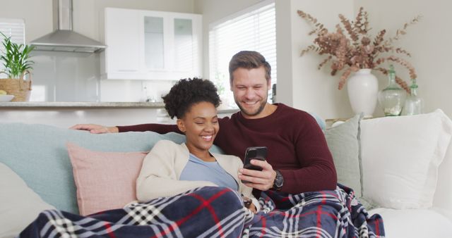 Image of happy diverse couple embracing and using smartphone on sofa. Love, relationship and spending quality time together at home.