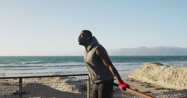 Man stretching his legs and leaning against a railing by the beach, ocean waves and mountains in background. Perfect for themes on fitness, outdoor exercise, morning routine, healthy lifestyle, and motivational content.