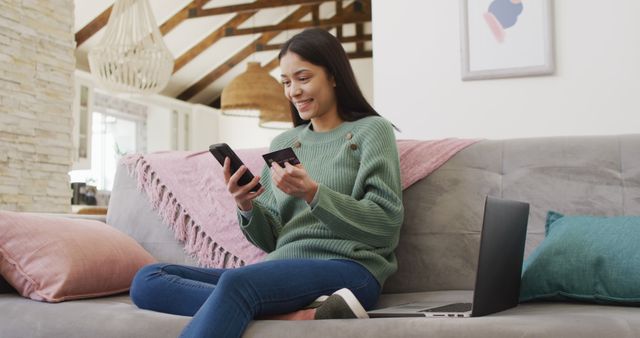 Young woman in a green sweater sitting comfortably on a sofa in a cozy living room, smiling while using a smartphone and holding a credit card. A laptop rests beside her, indicating a tech-savvy environment. Ideal for illustrating online shopping, digital transactions, modern lifestyle, and e-commerce concepts. Perfect for ads, blogs, and articles related to online shopping, digital marketing, and home-based business.