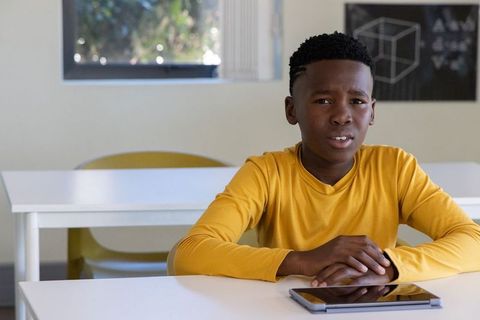 African american boy sitting at desk in mustard shirt holding tablet with confused look