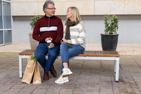 Middle-aged couple sitting on bench sharing coffee and smartphone in urban courtyard