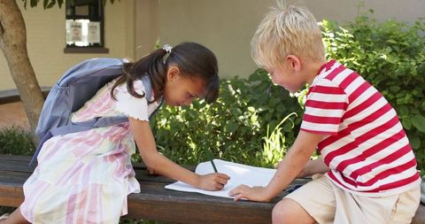 Children collaborating on homework outdoors writing in notebook on bench