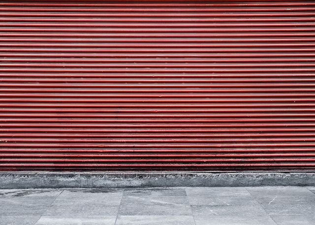 Red metal roller shutter covering entrance with concrete walkway in front. Ideal for use in urban and industrial themes, security concepts, street photography, or backgrounds for advertisements and architectural designs.