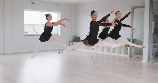 Ballet dancers dressed in black practicing jumps in a spacious studio with large window. Each dancer is in mid-air, displaying precision and grace. This image can be used for dance school promotions, fitness and flexibility articles, or inspiring posters about dedication and hard work.