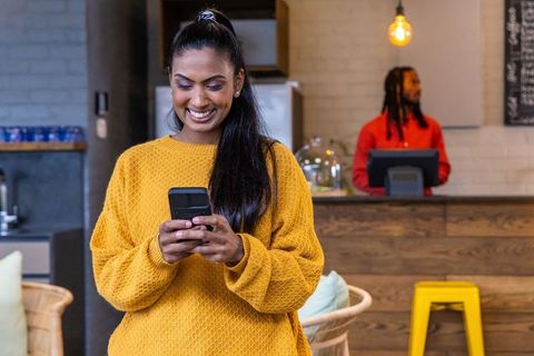 Indian woman texting with smartphone while smiling in cafe with african american barista
