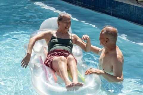 Senior asian couple enjoying sunny pool on inflatable lounger, smiling and holding hands