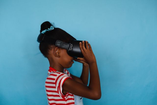 This image shows an African American elementary girl using VR glasses against a blue background. Ideal for use in educational materials, technology promotions, virtual reality content, and advertisements focusing on innovative learning methods.